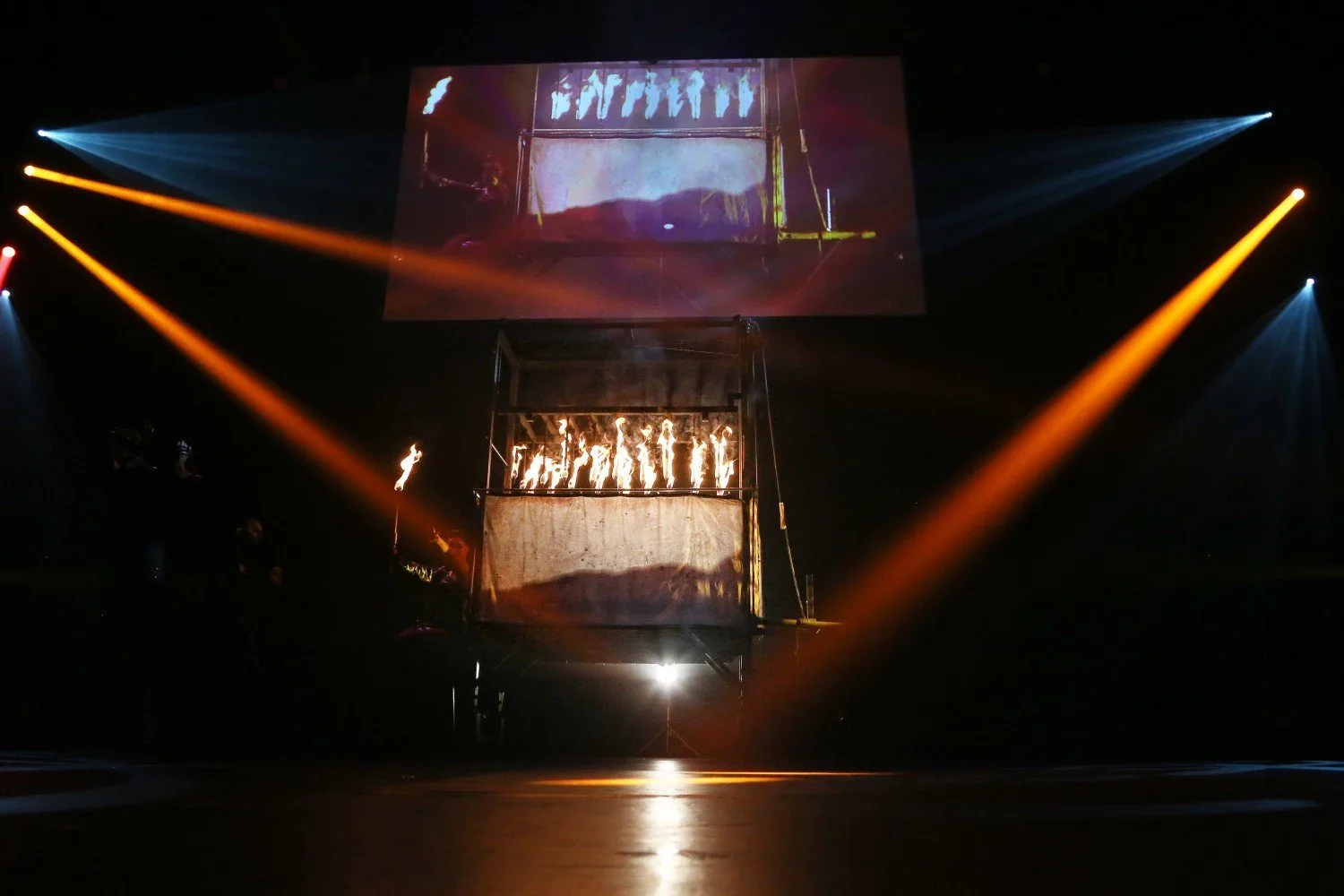 A suspended cage of flames lit by crossing beams on The Illusionists stage
