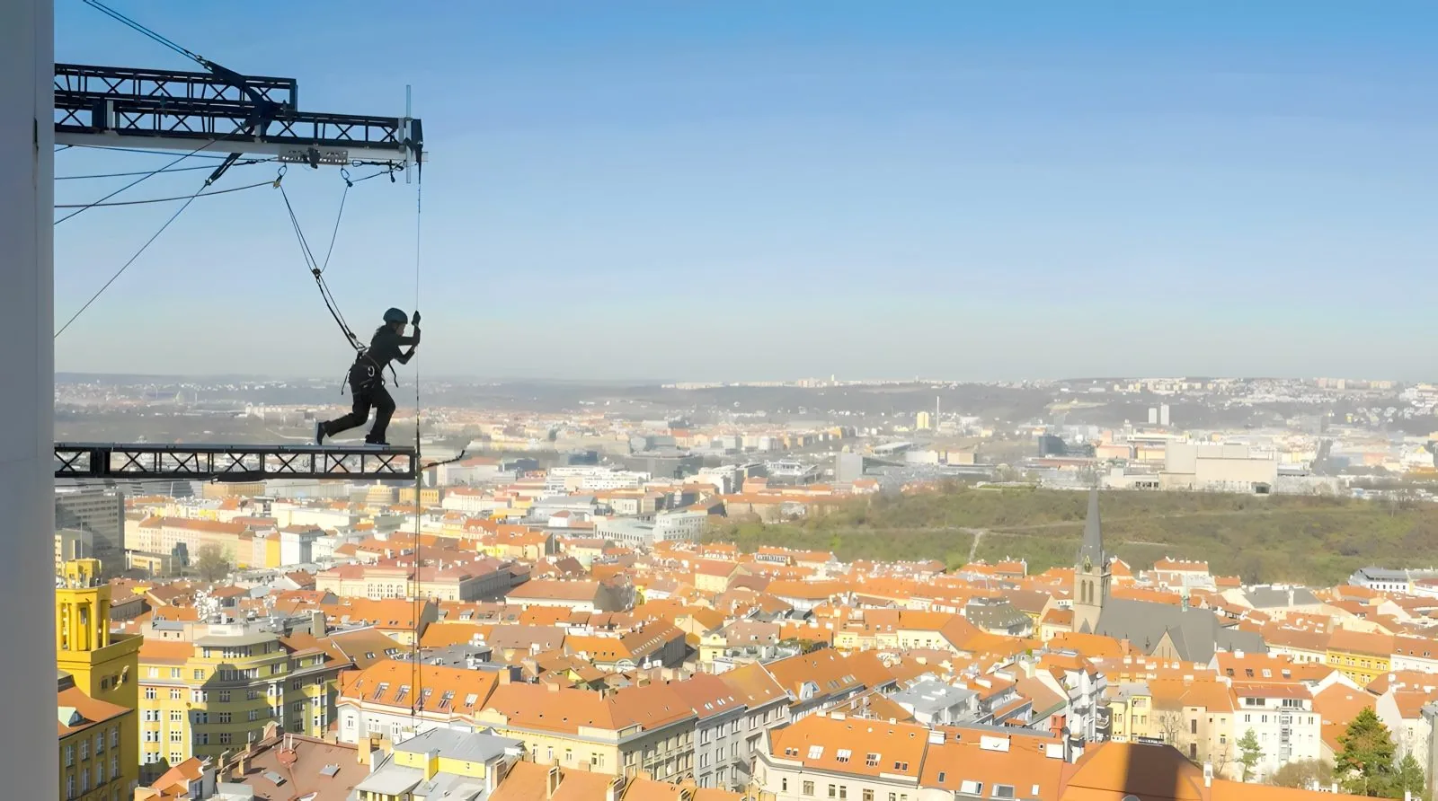 A harnessed racer on a truss platform off a TV tower, with Prague rooftops far below